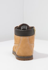 Tan suede nilkkurit, joissa on musta nahkakaulus, sivuvetoketju, valkoinen ommel ja teksturoitu kumipohja.