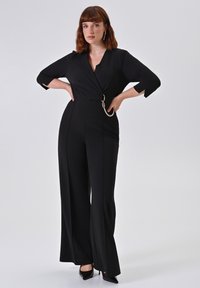 Jumpsuit nero con scollatura a V, maniche a tre quarti e gambe ampie. Presenta una catena decorativa in vita. Tessuto liscio.