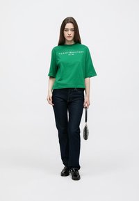 Zelena pamučna majica s kratkim rukavima i bijelim logotipom, kombinirana s tamnoplavim trapericama i crnim cipelama. Crna torbica s teksturiranim uzorkom.