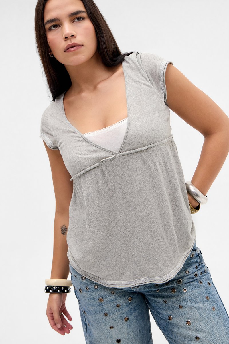 Νεαρή γυναίκα με μακριά σκούρα μαλλιά που φοράει γκρι κοντομάνικο τοπ πάνω από λευκό φανελάκι, τζιν με τρουκς και πολλαπλά βραχιόλια.