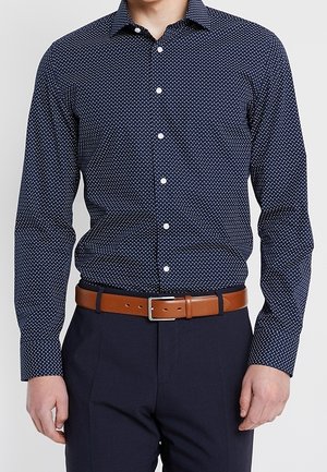 Mann trägt ein dunkelblau gemustertes Hemd mit Knöpfen, dunkle Hose und einen braunen Leder Gürtel mit silberner Schnalle.