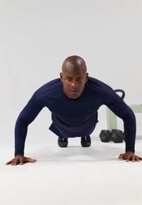 Tengerészeti hosszú ujjú pólót viselő sportoló fekvőtámaszokat végez egy fehér felületen, a háttérben kézi súlyzók és egy kettlebell található.
