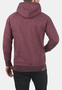 Sweatshirt à capuche bordeaux avec un tissu texturé, des manches raglan et un ourlet côtelé. Présente une petite étiquette de logo en bas à gauche.