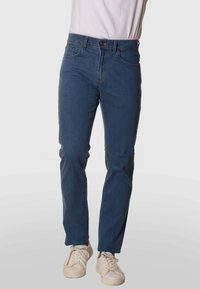 Jeans de mezclilla azul oscuro con corte recto, cinco bolsillos, cierre de cremallera y trabillas para el cinturón, combinados con zapatillas blancas sobre un fondo sencillo.