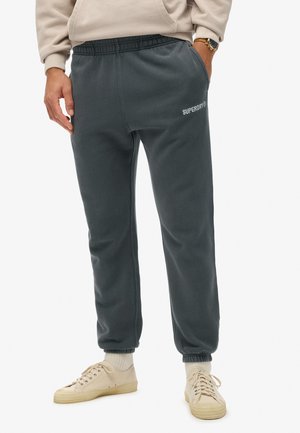 Grijze joggingsbroek van zacht materiaal, met een trekkoord in de taille, zijzakken en het witte "SUPERDRY" logo op de linker bovenbeen.
