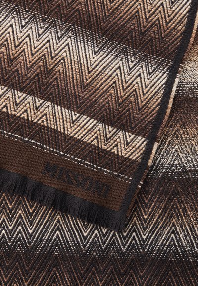 Vævet tæppe med et zigzag-mønster i brune og creme farver, med en frynset kant og et præget logo på en sort label.