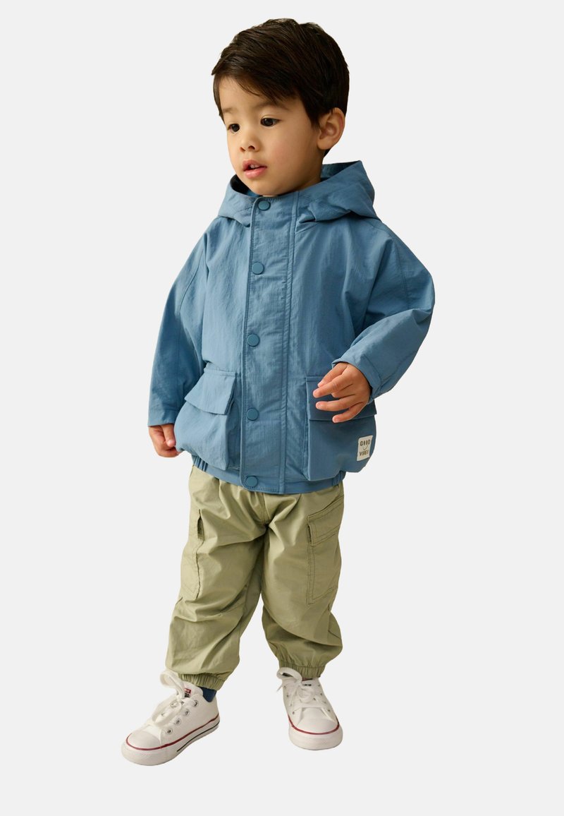 Jeune enfant portant une veste à capuche bleue, un pantalon cargo beige et des baskets blanches, debout et regardant sur le côté.