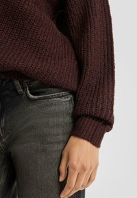 Bordowy sweter dziergany o fakturowanym wzorze, z prążkowanymi mankietami i luźnym krojem, zestawiony z ciemnoszarymi dżinsami. Zbliżenie na rękaw i dłoń.