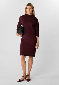 Donna che indossa un vestito bordeaux lungo fino al ginocchio con maniche a tre quarti, tiene in mano una borsa nera e indossa tacchi slingback bordeaux.