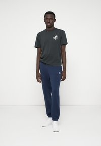 Tamsiai pilkas marškinėliai su spausdintu dizainu ant krūtinės, derinami su tamsiai mėlynomis sportinėmis kelnių. Abu drabužiai turi laisvą siluetą. Baltos sportbačiai užbaigia įvaizdį.