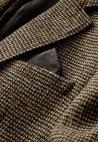 Tweed blazer in brown with blue and green flecks. Features a notch lapel and a textured pocket square in dark brown with a patterned design.