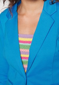 Modrý blazer s texturovaným povrchem, nošený přes vícestupňový háčkovaný top s pruhy v růžové, žluté, zelené a modré barvě.