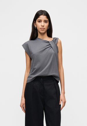 Kvinne med langt mørkt hår som har på seg en ermeløs grå topp med en knutedetalj på den ene skulderen og svarte høytlivsbukser.