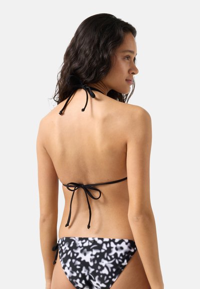 Junge Frau mit schwarzen, welligen Haaren, die einen schwarz-weiß gemusterten Bikini trägt, von hinten gesehen, sichtbarer Neckholder-Oberteil mit Bindung und seitlich gebundene Bikinislips.