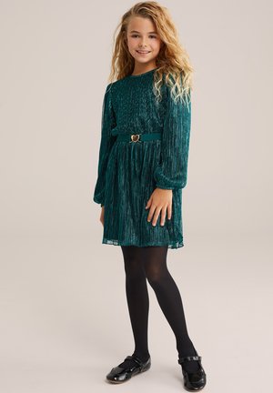Rochie plisată verde cu mâneci lungi, țesătură texturată, curea cu cataramă în formă de inimă, asortată cu colanți negri și pantofi negri lucioși.