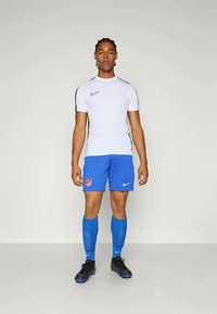 Atleta de pie, de frente, vistiendo una camiseta blanca de Nike, pantalones cortos azules del Atlético de Madrid y calcetas, y botas de fútbol negras, sobre un fondo gris.