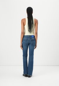 Blå denim flared jeans med medium vask, baklommer og et blått logo-patch på livet, paret med en lys gul tanktop.