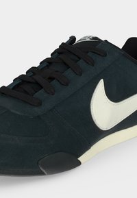 Μαύρο sneaker Nike με λευκό λογότυπο Swoosh, μαύρα κορδόνια και υφασμένο πάτο, σε φωτογραφία από κοντινή πλευρική γωνία.