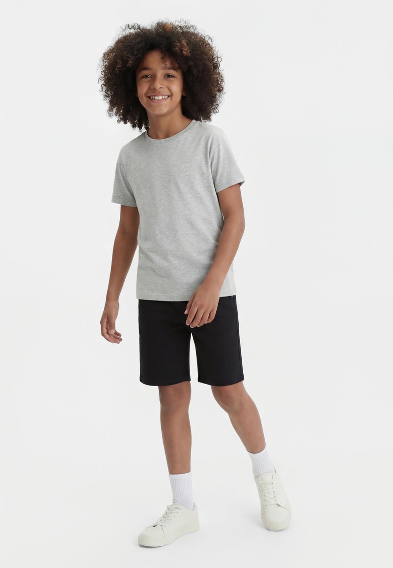 Enfant souriant aux cheveux bouclés debout, portant un t-shirt gris clair, un short noir, des chaussettes blanches et des baskets blanches sur un fond blanc.