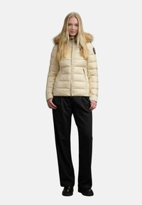 Giacca puffer di un beige chiaro con cappuccio in finto pelo, chiusura con zip e due tasche laterali. Indossata con pantaloni neri larghi e scarpe nere.
