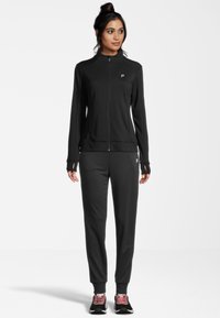 Tuta sportiva nera con giacca zip-up e pantaloni. Il tessuto appare liscio; la giacca presenta aperture per i pollici e un piccolo logo. Le scarpe hanno dettagli rosa.