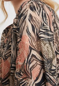 Blouse transparente avec imprimé animal multicolore dans des tons beige, noir et corail. Dispose d'un tissu léger et texturé avec une subtile étincelle.