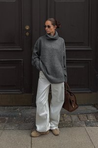 Suéter gris de gran tamaño de punto con un cuello alto, combinado con unos pantalones anchos blancos y calzado beige. Bolso marrón con detalles de hardware.
