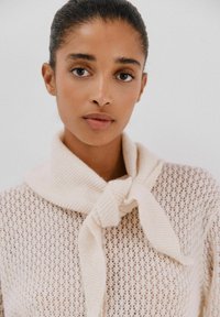 Maglione lavorato a maglia in crema con design a rete aperta, dotato di un ampio colleto a scialle annodato nella parte anteriore. La superficie liscia mette in risalto i dettagli.