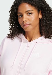Lyse rosa zip-up hettegenser med myk tekstur, snøring på hetten og hvit logo på brystet. Nærbilde av halslinjen og ansiktet.
