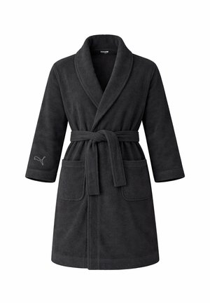 Lächelnde Frau, die einen schwarzen knielangen Mantel mit Gürtel und Taschen trägt, steht vor einem neutralen Hintergrund mit einer Hand in der Tasche.