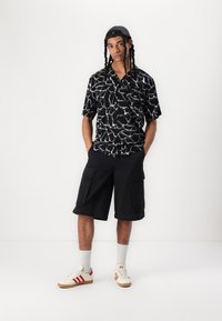 Camicetta nera a maniche corte con motivo astratto bianco, abbinata a pantaloni cargo neri fino al ginocchio. Completato con sneakers crema e calzini grigi.