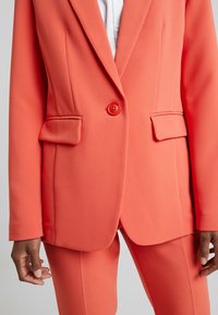 Koralowa marynarka z gładkiej tkaniny z zapięciem na pojedynczy guzik, dwoma przednimi kieszeniami, klapami z wycięciem i dopasowanym krojem. Stylizowana z dopasowanymi spodniami.