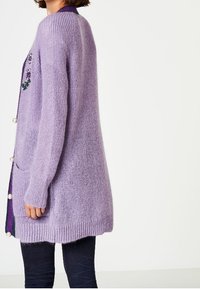 Lavendelstickad cardigan med långa ärmar, rundad nederkant ochpärlprydnader på knapparna. Har ett texturerat, mjukt material.