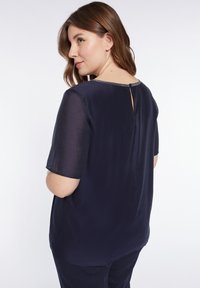 Blusa blu navy a maniche corte in tessuto leggero, con un dettaglio a apertura a chiave sulla schiena e un sottile bordo decorativo attorno al collo.