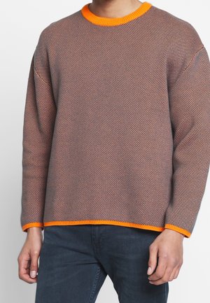 Brauner, strukturierter Langarm-Pullover mit leuchtend orangefarbenem Besatz an Kragen, Bündchen und Saum, getragen mit dunkler Hose, Oberkörper sichtbar.