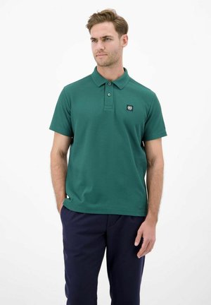 Mann trägt grünes kurzärmeliges Poloshirt mit Knöpfen und dunkelblaue Hose, steht mit einer Hand in der Tasche vor weißem Hintergrund.