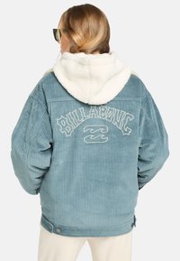 Lys blå cordjakke med hvid fleeceforet hætte, prydet med broderet "Billabong" logo på ryggen. Trykknapper ved kanten.