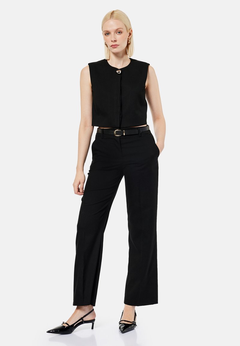 Woman with blonde hair wearing a sleeveless black cropped top, black belted trousers, and black pointed-toe heels, standing with one hand in pocket.