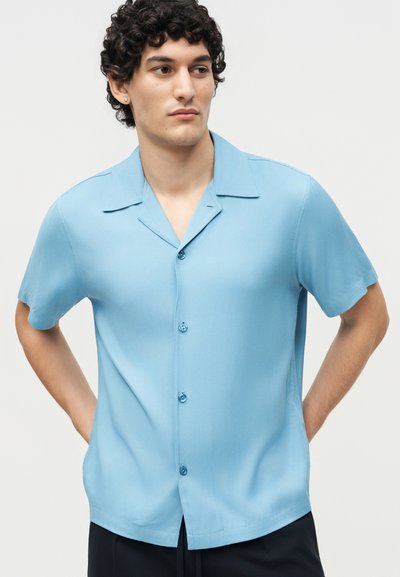 Νέος άνδρας με σγουρά σκούρα μαλλιά που φορά ένα ανοιχτό μπλε πουκάμισο με κοντά μανίκια, στέκεται με τα χέρια πίσω από την πλάτη και κοιτάζει αριστερά.