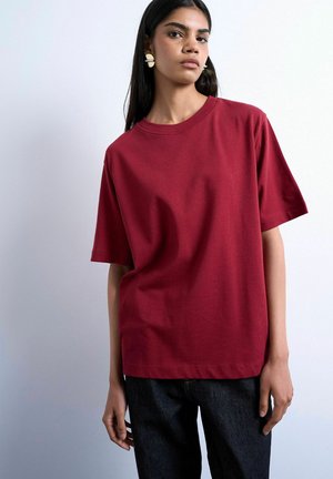Femme aux longs cheveux noirs portant un t-shirt rouge uni à manches courtes et un pantalon noir, debout devant un fond clair uni.