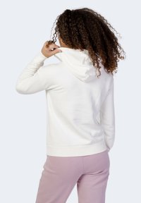 Felpa bianca con cappuccio, polsini e orlo a coste. Il tessuto sembra morbido. Indossata con pantaloni della tuta rosa chiaro.
