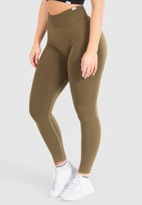 Leggings olive verdi a vita alta realizzati in materiale elasticizzato con una superficie liscia, dotati di una cucitura laterale a contrasto e abbinati a scarpe da ginnastica bianche.