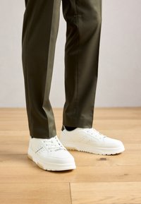 Persoană îmbrăcată cu pantaloni verzi măslinii și adidași albi Bugatti, stând pe un podea din lemn.