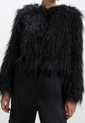 Person, die eine langärmelige, schwarze, zottelige Kunstfelljacke und schwarze Hose trägt, von den Schultern bis zu den Oberschenkeln vor einfarbigem Hintergrund gezeigt.