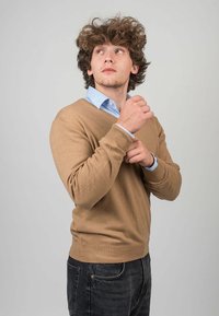 Ruskea neulepaita V-pääntiellä, yhdistettynä vaalea siniseen kauluspaitaan. Malli säätää neulepaidan hihoja. Tummat farkut täydentävät asun.