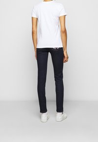 Osoba v bílém tričku, tmavých úzkých džínách srdcového vzoru na pravé zadní kapse, bílých ponožkách a bílých teniskách, stojící zády.