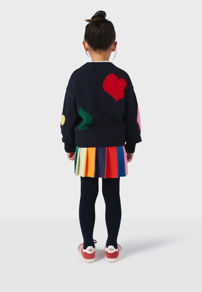 Kind trägt einen marineblauen Pullover mit bunten Formen, einen plissierten, mehrfarbigen Rock, marineblaue Strumpfhosen und rote Schuhe mit weißen Akzenten.