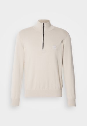 Ljust beige tröja med kvartdragkedja, stickad textur och en liten logotyp på bröstet. Åtsittande mudd och ribbad midja.
