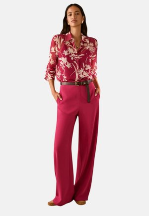 Vrouw draagt een rode bloemen lange mouwen blouse, ingestopt in een wijde rode broek met een zwarte riem, staand met handen in de zakken.