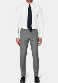 Camicia bianca con colletto, abbinata a pantaloni grigi a scacchi e una cravatta blu scuro; le scarpe sono in pelle nera. Taglio su misura.